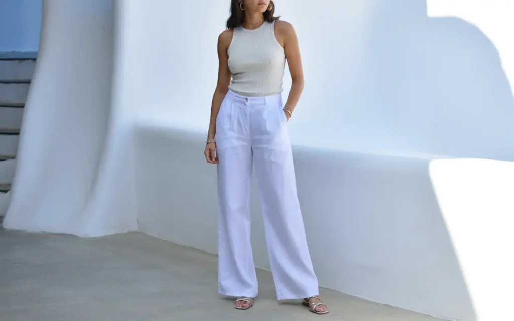  Woman in crisp white wide-leg linen trousers and a fitted ivory ribbed tank top with strappy flat sandals and gold jewelry standing against a whitewashed wall