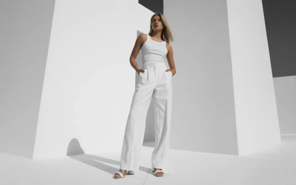  Woman wearing a full all-white summer outfit including wide-leg trousers, a fitted tank top, and heeled sandals photographed against a minimalist white architectural backdrop in bright midday light
