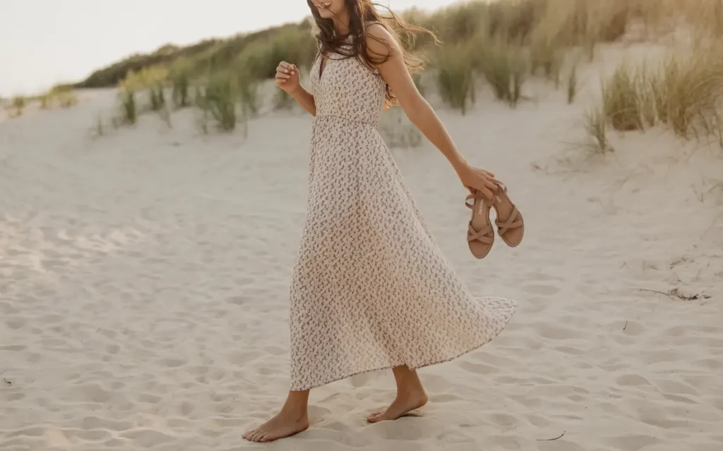 Woman in a flowy ditsy floral maxi dress in soft muted tones walking barefoot on a sandy beach path holding flat leather sandals with golden light